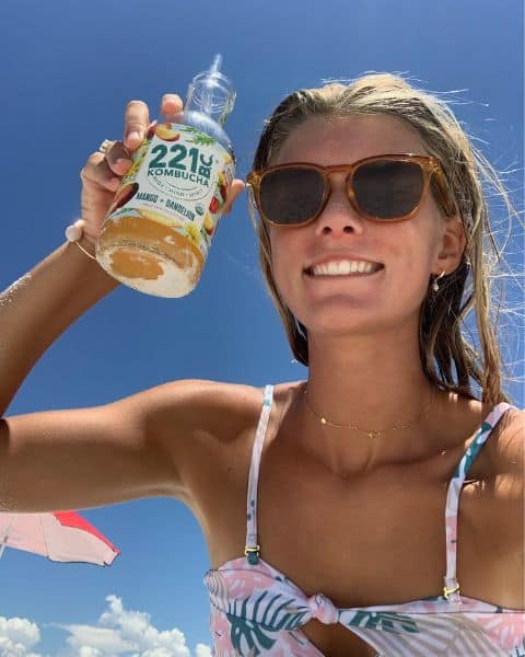 woman in swimsuit on the beach holding up a bottle of mango + dandelion kombucha flavor