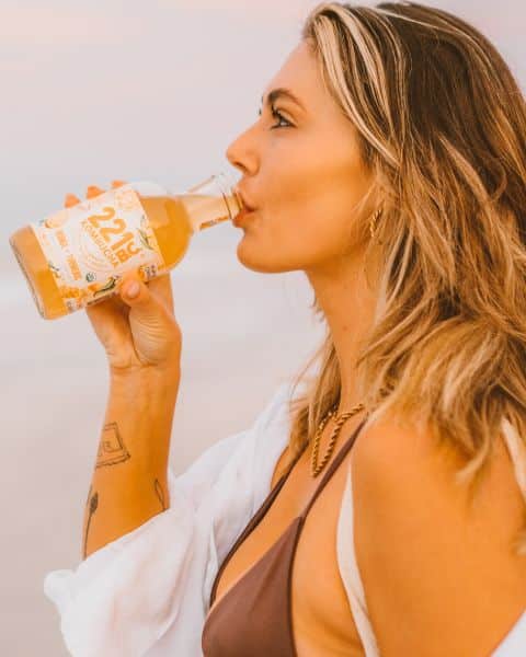 woman in bikini drinking a bottle of orange + turmeric kombucha flavor